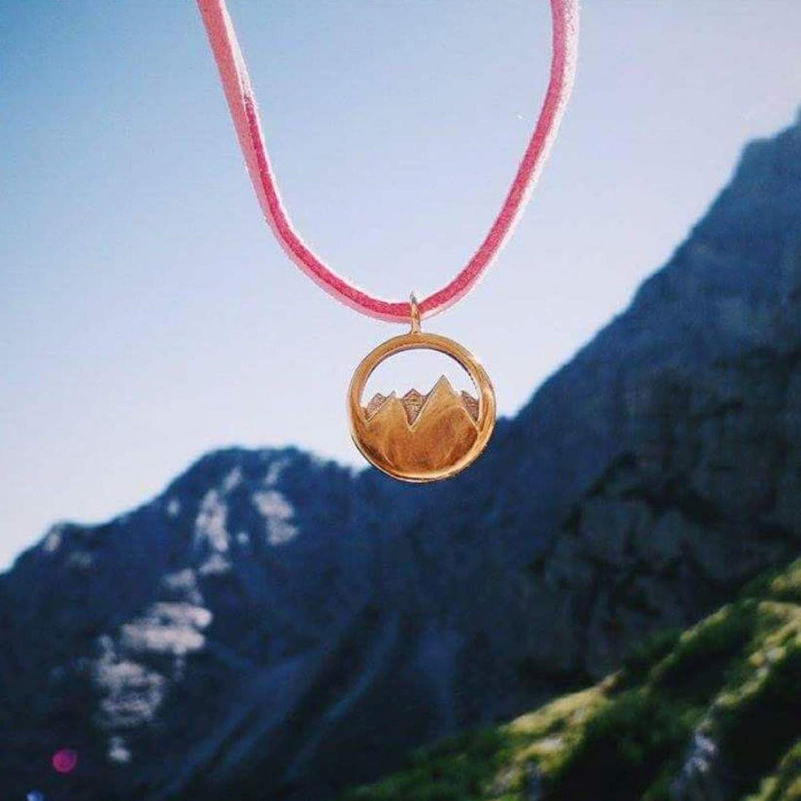 Der Silver+Surf Silber Schmuck Berg 3D vergoldet Anhänger - ein rundes, vergoldetes Stück aus 925er Sterlingsilber mit einem Bergmotiv - hängt an einer rosafarbenen Kordel vor einem verschwommenen Hintergrund aus zerklüfteten Bergen.