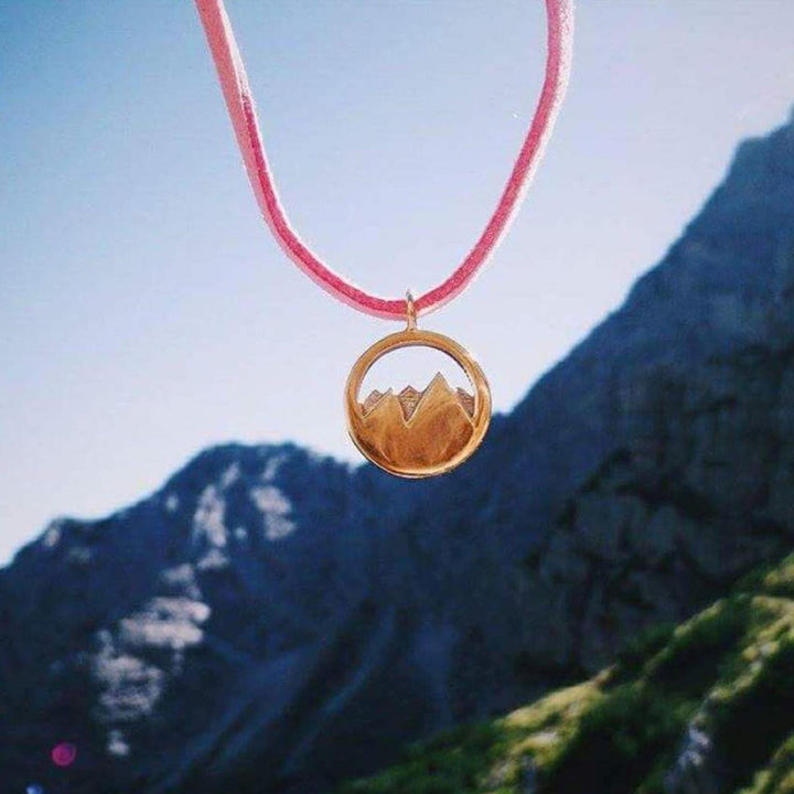 Der Silver+Surf Silber Schmuck Berg 3D vergoldet Anhänger - ein rundes, vergoldetes Stück aus 925er Sterlingsilber mit einem Bergmotiv - hängt an einer rosafarbenen Kordel vor einem verschwommenen Hintergrund aus zerklüfteten Bergen.