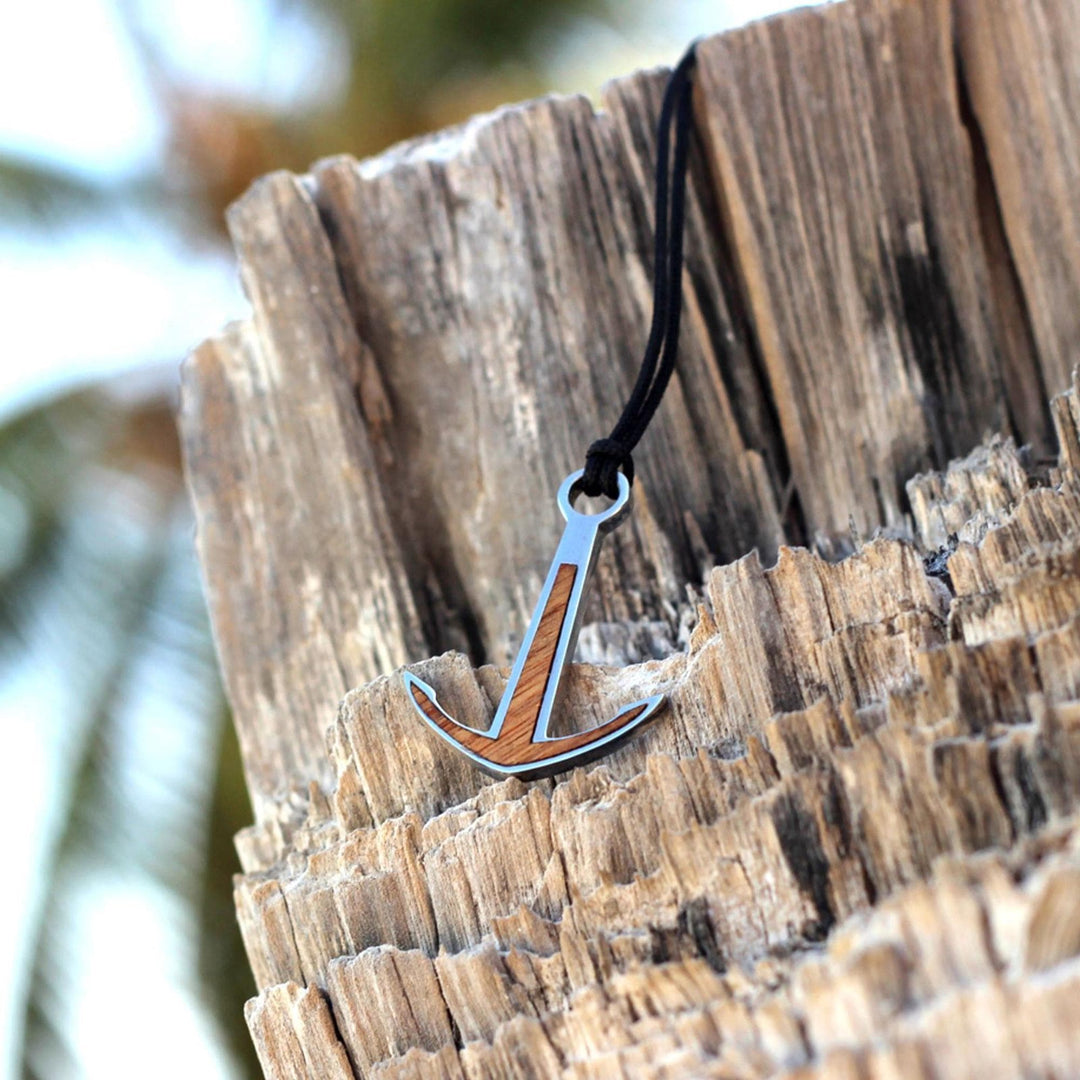 Das Silver+Surf Silber Schmuck BootAnker Wood Halsband der Marke Silver+Surf liegt an einer schwarzen Kordel auf verwittertem Holz, unscharfe Palmblätter bilden den Hintergrund.