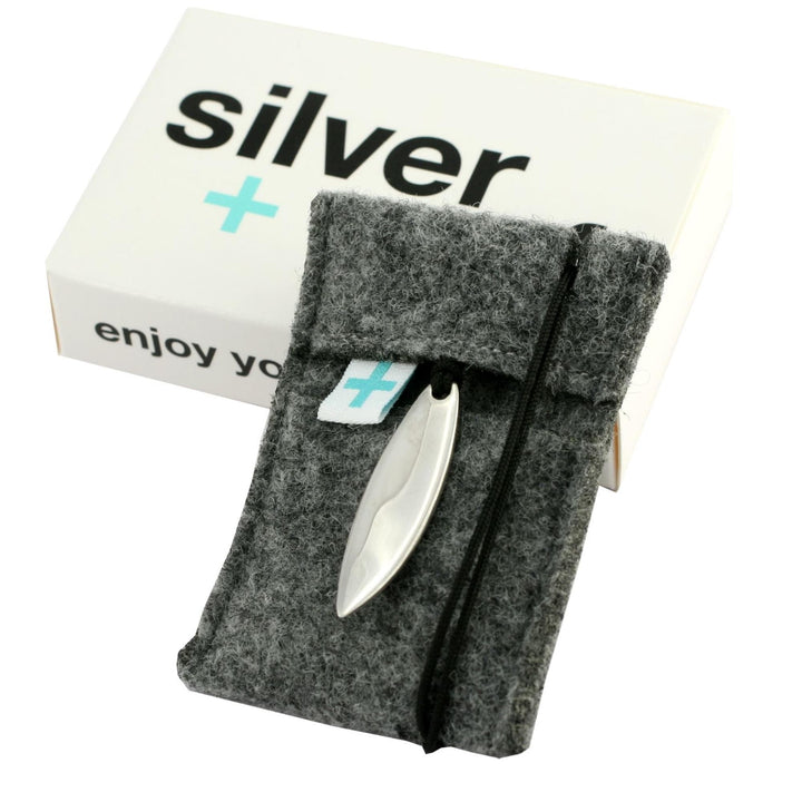 Hinter einem grauen Filzbeutel mit schwarzer Kordel befindet sich eine weiße rechteckige Schachtel mit der Aufschrift Silver+Surf. Am Beutel befestigt ist ein glänzender BootAnker Wood Halsband-Silber Schmuck von Silver+Surf, handgemacht in Europa. Auf der Schachtel steht außerdem "enjoy yo.