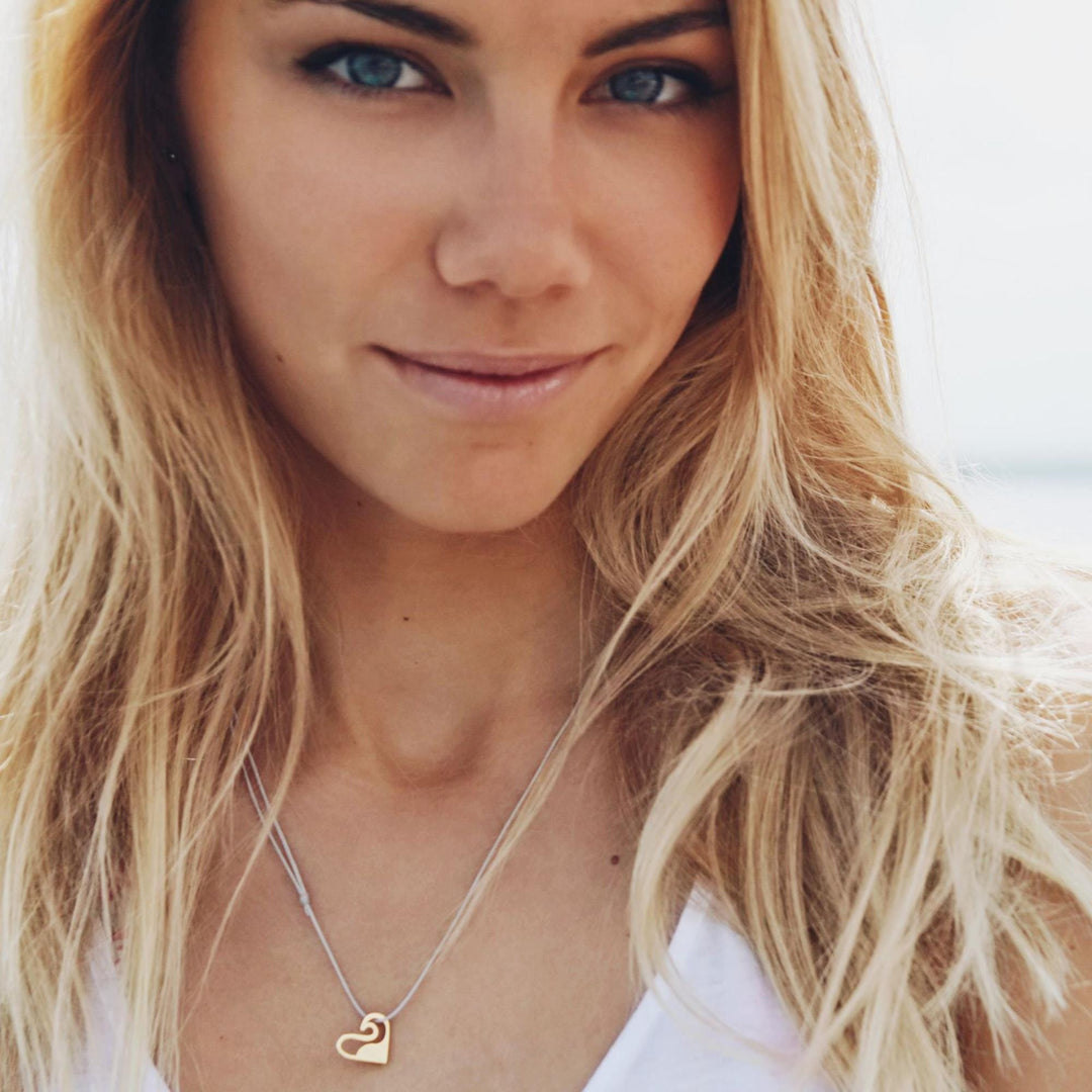 Eine Frau mit langen blonden Haaren und blauen Augen trägt ein weißes Oberteil und die goldene Halskette Silver+Surf Silber Schmuck Love Welle Wave. Sie steht im Freien und schaut mit einem leichten Lächeln in die Kamera.