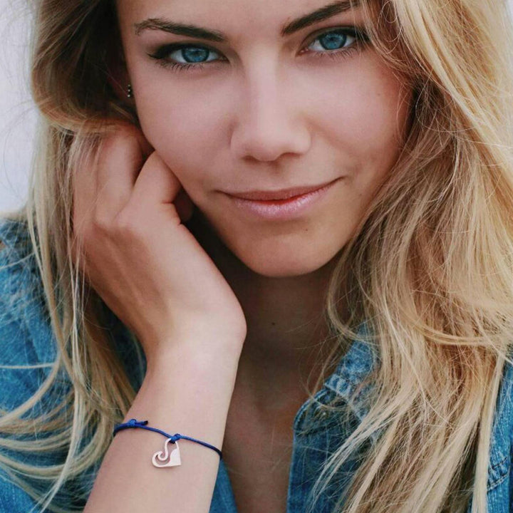 Eine Frau mit langen blonden Haaren und blauen Augen trägt ein Jeanshemd und das goldene Armband Silver+Surf Love Wave, stützt ihre Hand auf ihr Gesicht und schaut mit einem leichten Lächeln in die Kamera.