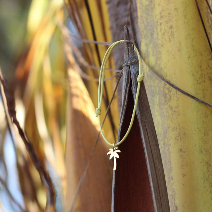 Das Silver+Surf Silber Schmuck Palme vergoldet, ein gelbes Kordelarmband mit einem vergoldeten Palmenanhänger, hängt an einer braunen Palmwedelspitze mit verschwommenen gelben und braunen Palmenblättern im Hintergrund.