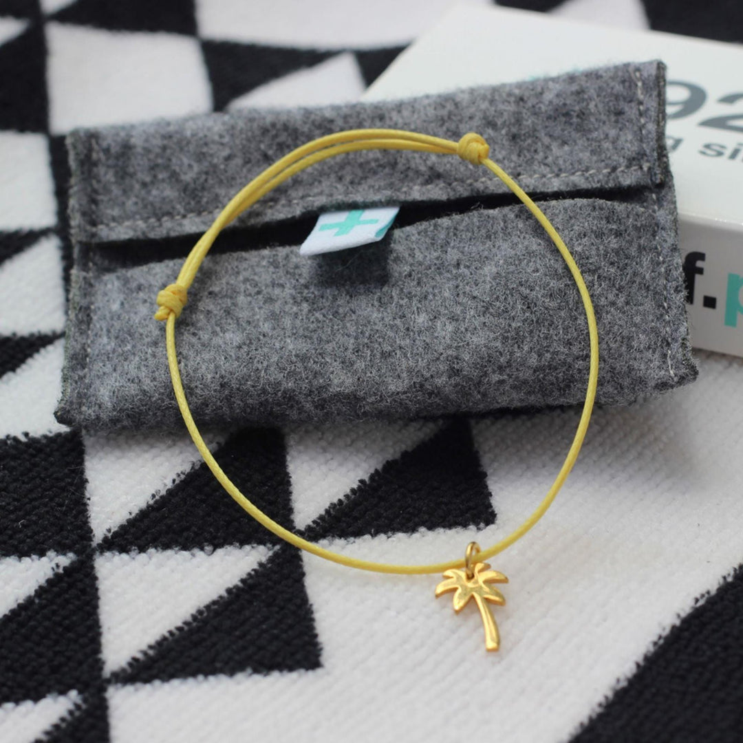 Ein gelbes Kordelarmband mit einem vergoldeten Palmenanhänger von Silver+Surf ruht auf einem grauen Filzbeutel. Dahinter befindet sich eine weiße Silver+Surf-Box mit schwarzem Text, die auf gemustertem schwarz-weißem Stoff liegt.