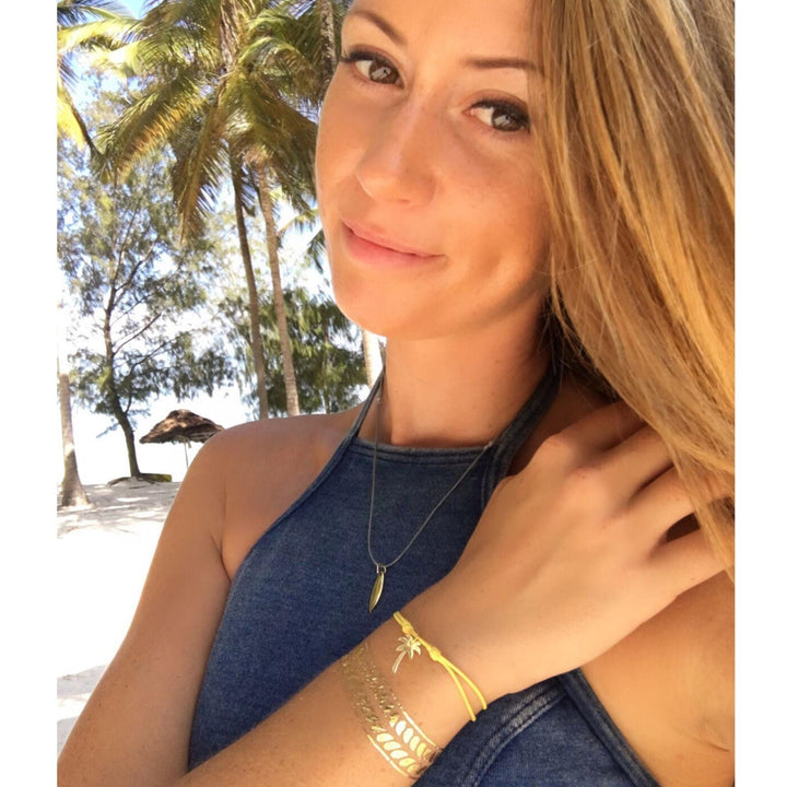 Eine Frau in einem blauen ärmellosen Oberteil und mit Goldschmuck, darunter die Silver+Surf Silber Schmuck Palme vergoldet, steht im Freien mit Palmen und einem Sandstrand im Hintergrund und lächelt leicht in die Kamera.