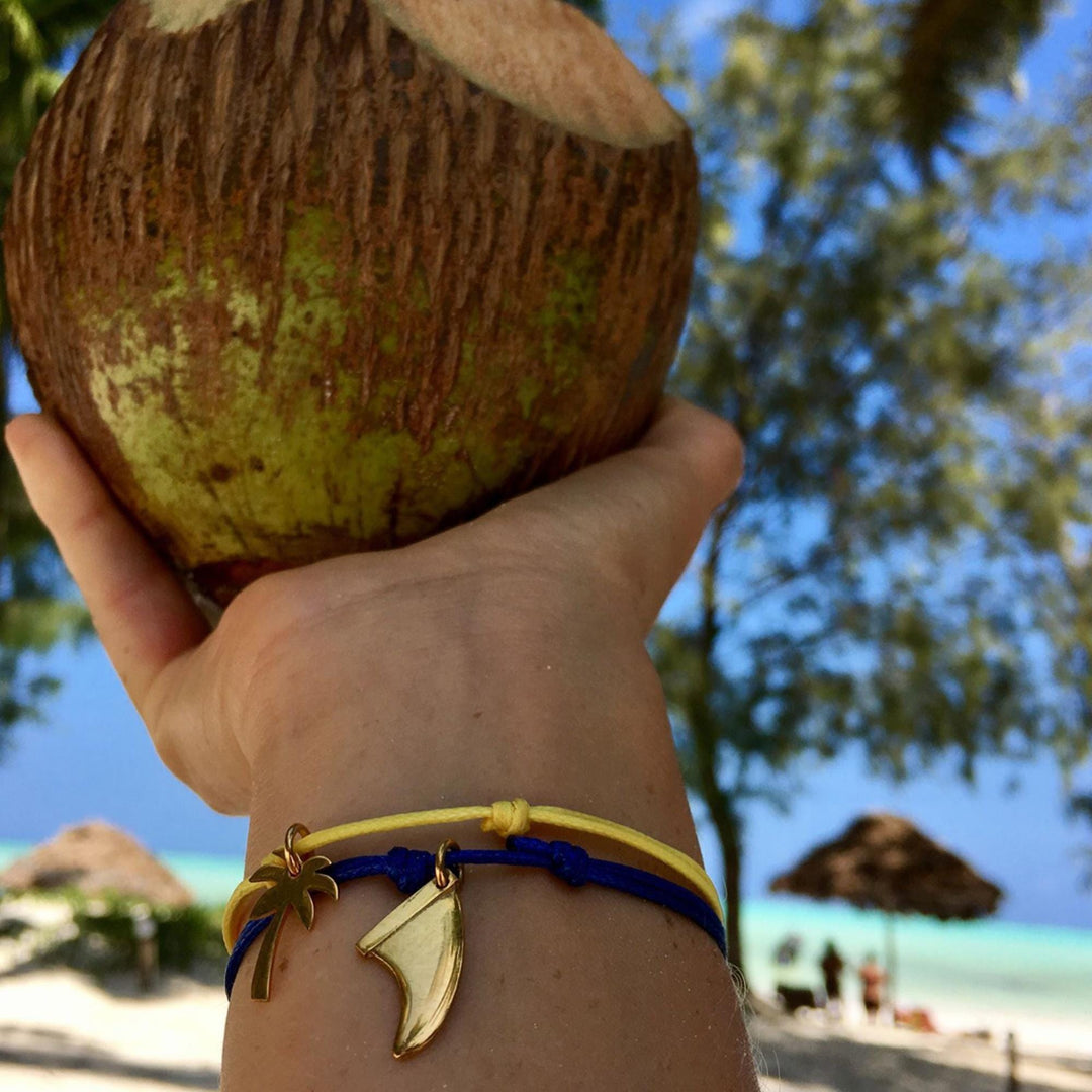 Eine mit Silver+Surf Silber Schmuck Palme vergoldet und anderen goldenen Armbändern geschmückte Hand hält eine Kokosnuss an einem tropischen Strand, mit Palmen, Meer und strohgedeckten Sonnenschirmen im Hintergrund.