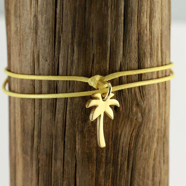 Ein gelbes Kordelarmband von Silver+Surf mit einem kleinen vergoldeten Palmen-Anhänger (Silber Schmuck Palme vergoldet) ist um ein Stück verwittertes Holz gewickelt.