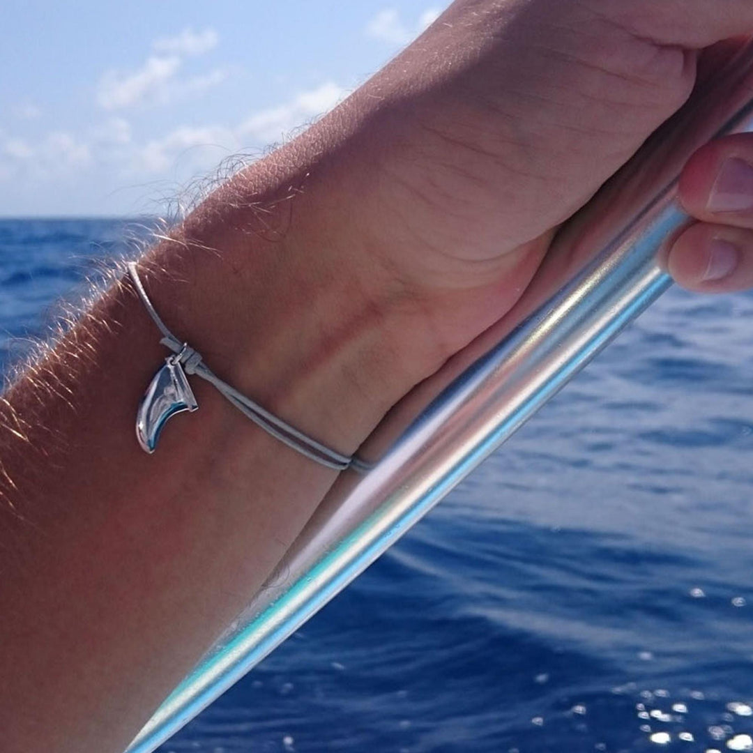 Eine Hand hält eine Metallstange auf einem Boot mit Meer und Himmel im Hintergrund. Sie trägt das Silver+Surf Silber Schmuck Surfboard Finne Armband von Silver+Surf.