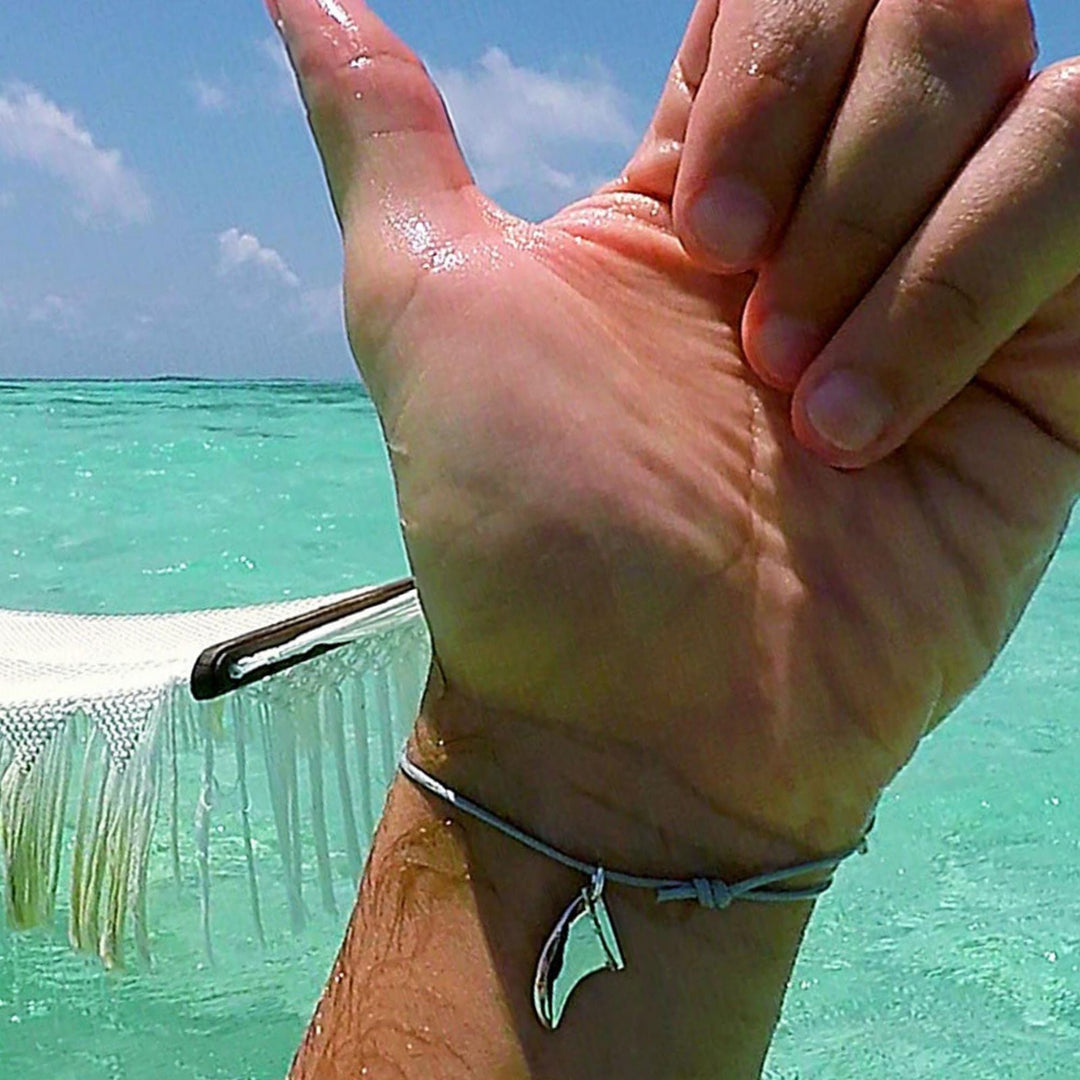 Eine Nahaufnahme einer Hand, die das Silver+Surf Silber Schmuck Surfboard Finne Armband trägt, vor türkisfarbenem Ozeanwasser und einer weißen Hängematte mit einem meist klaren, leicht bewölkten Himmel.