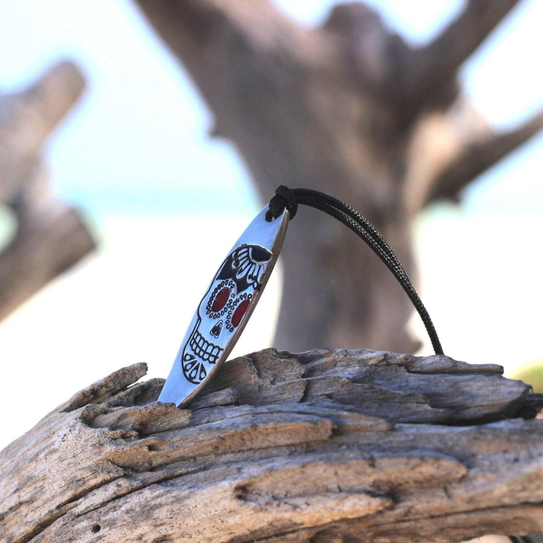 Ein Silver+Surf Silber Schmuck Surfboard mit Totenkopf Anhänger ruht auf Treibholz. Das gemalte Totenkopfmotiv auf einer schwarzen Kordel hebt sich von verschwommenen Ästen und einem Strand ab und fängt den Küstengeist der Marke ein.