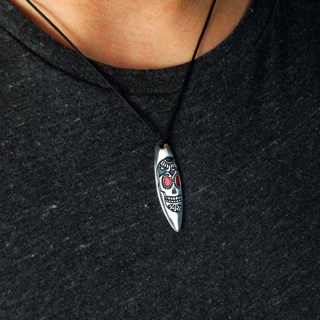 Jemand in einem schwarzen T-Shirt trägt eine Silver+Surf-Halskette mit einer schwarzen Kordel und einem 925er Sterling-Silber-Surfbrett-Anhänger, der einen Totenkopf mit roten Augen zeigt.