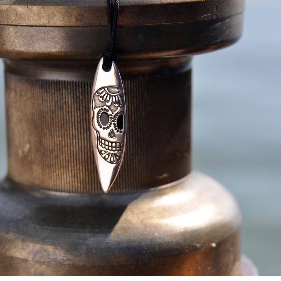Das Silver+Surf Silber Schmuck Surfboard mit Totenkopf von Silver+Surf aus 925 Sterling Silber hängt an einer schwarzen Kordel vor unscharfem Hintergrund.