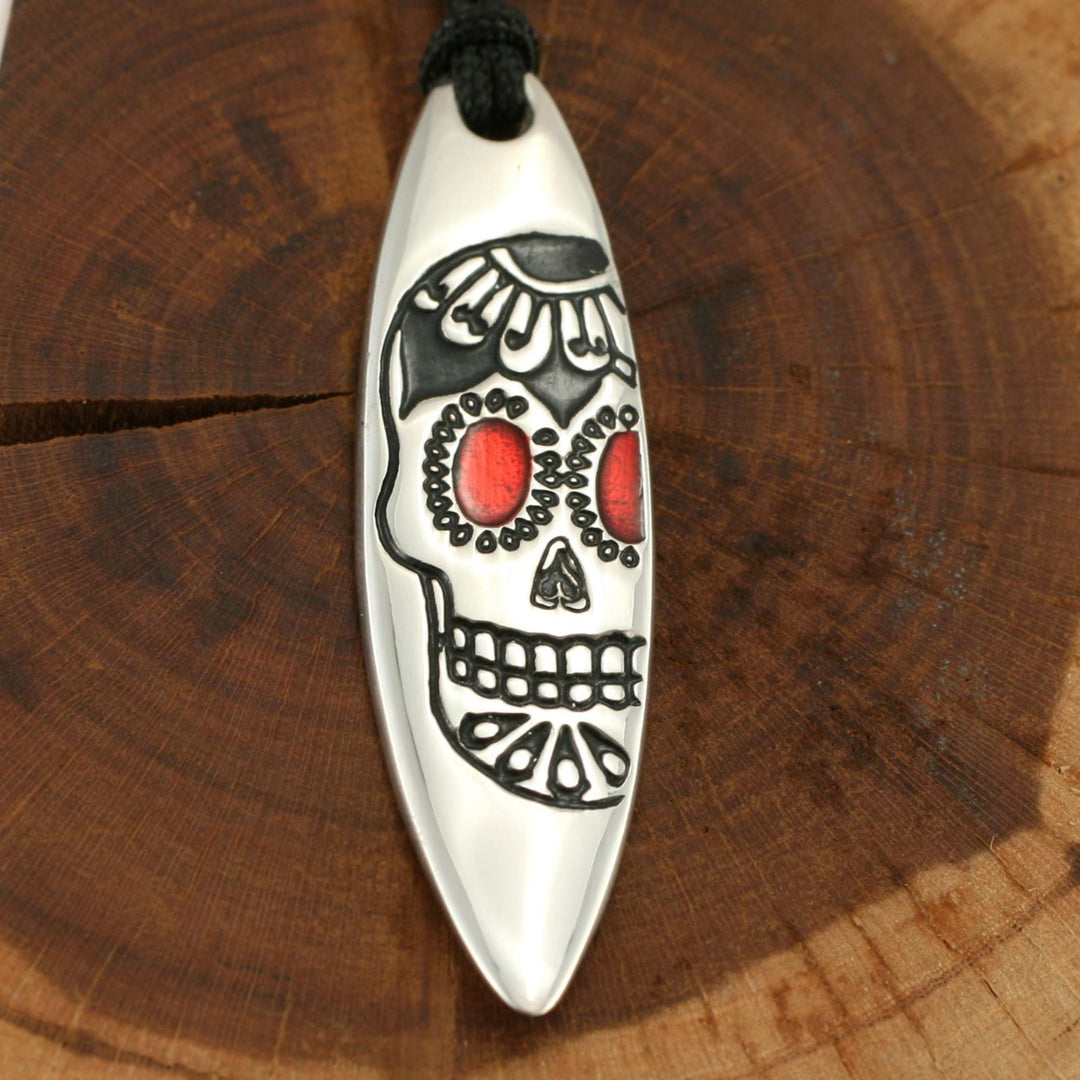 Der Silver+Surf Silber Schmuck Surfboard mit Totenkopf ist ein einzigartiger 925 Sterling Silber Anhänger mit einem Totenkopf-Design auf einer schwarzen Kordel, die auf einer runden Holzoberfläche angebracht ist - perfekt für jede Gelegenheit.