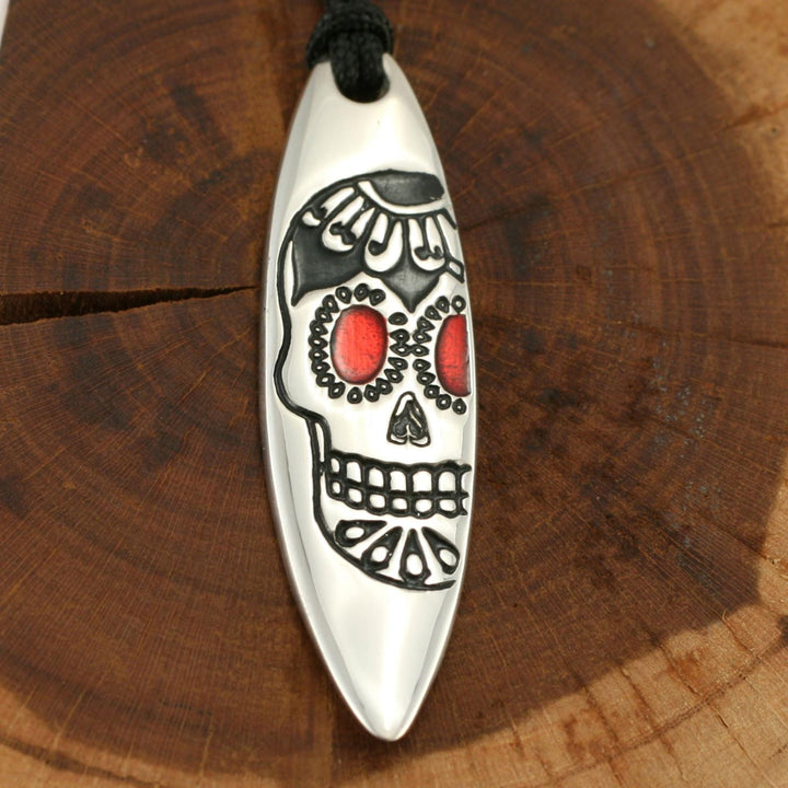 Der Silver+Surf Silber Schmuck Surfboard mit Totenkopf ist ein einzigartiger 925 Sterling Silber Anhänger mit einem Totenkopf-Design auf einer schwarzen Kordel, die auf einer runden Holzoberfläche angebracht ist - perfekt für jede Gelegenheit.