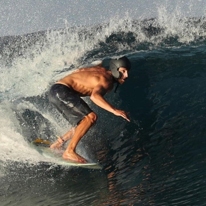 Ein Mann, der einen SIMBA Surf Wassersport Helm Sentinel von Simba Surf und schwarze Shorts trägt, surft auf einer Welle und lehnt sich auf seinem Brett nach vorne, während das Wasser um ihn herum spritzt.