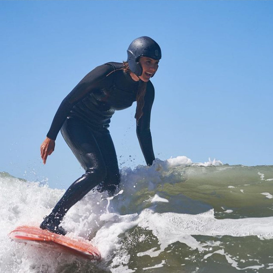 Ein Surfer in einem schwarzen Neoprenanzug und einem SIMBA Surf Wassersport Helm Sentinel von Simba Surf reitet auf einem roten Surfbrett auf einer kleinen Welle und balanciert mit gebeugten Knien und ausgestreckten Armen unter einem klaren blauen Himmel.