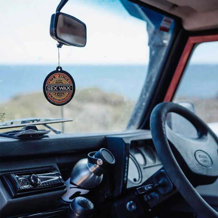 Blick in ein Autoinnere mit einem Lenkrad, einem Becherhalter mit einer Thermoskanne und einem SEX WAX Lufterfrischer von SEX WAX, der am Rückspiegel hängt. Der Duft von Surf Duft erfüllt die Luft, während das Meer und der Himmel durch das Fenster zu sehen sind.