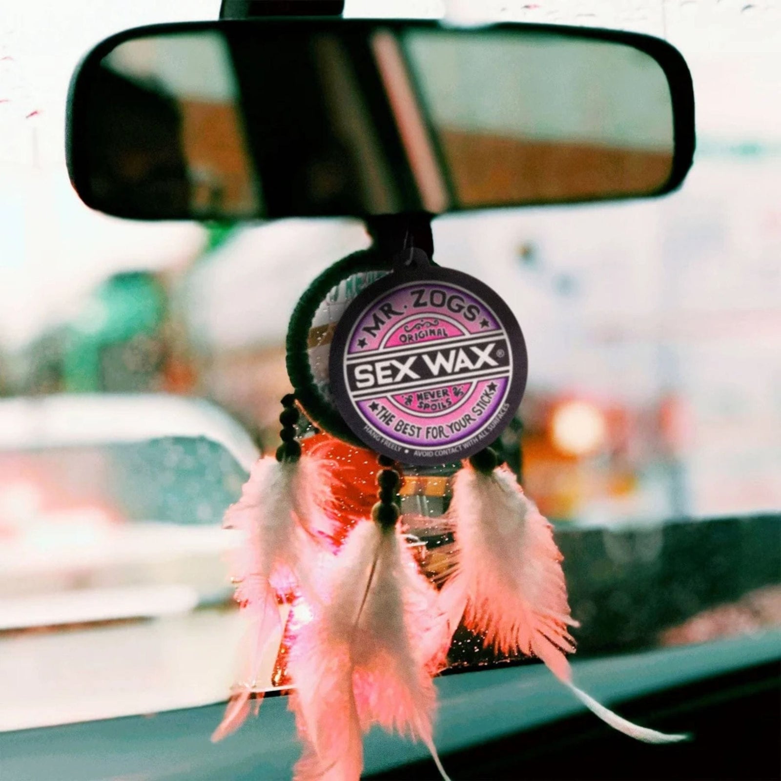 Ein SEX WAX Air Freshener Lufterfrischer von SEX WAX hängt mit rosa und weißen Federn geschmückt am Rückspiegel eines Autos, während Regentropfen auf der Scheibe die Sicht auf den Verkehr draußen trüben.