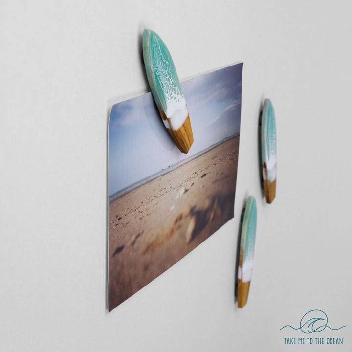 Ein Sandstrand-Foto wird an einer weißen Wand mit drei handgefertigten Surfboard-Magneten von Takemetotheocean festgehalten, die jeweils türkise, weiße und hölzerne Spitzen aufweisen. Das nachhaltige Ocean Art Logo erscheint in der unteren rechten Ecke.