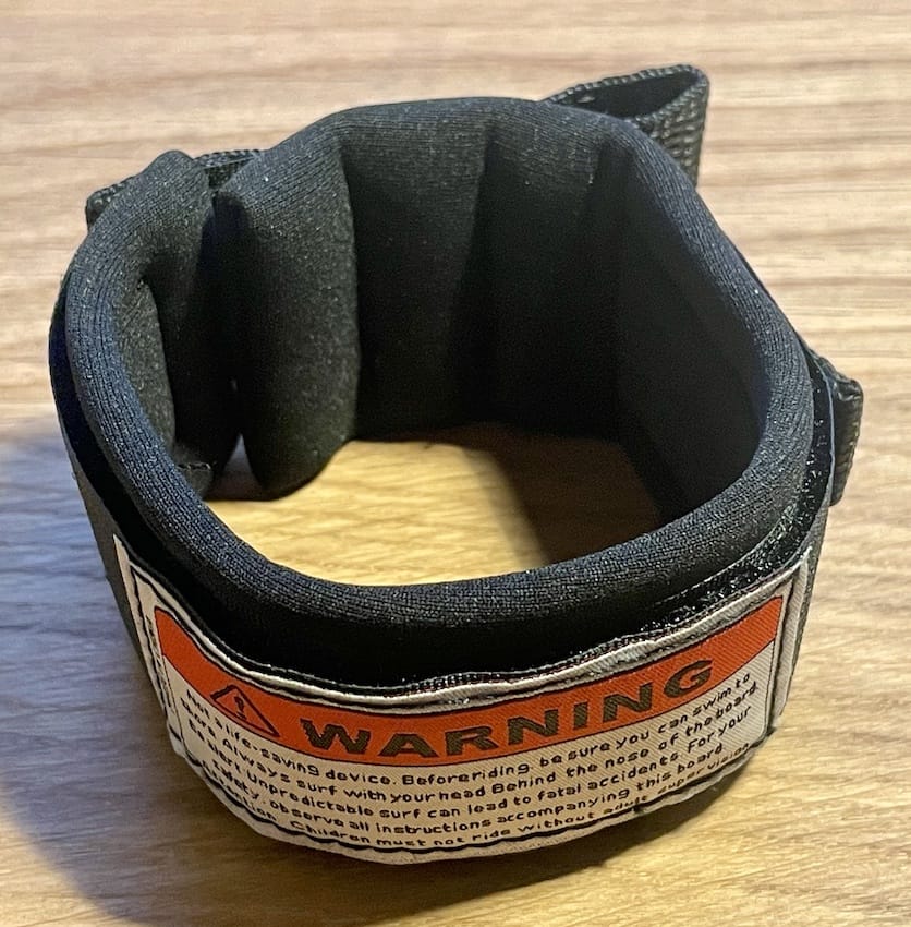 Ein gepolsterter schwarzer Klett-Fußfessel (Ersatzteil) von Rapid Surf Shop mit einem orange-weißen Warnschild mit Sicherheitshinweisen in kleiner Schrift auf einer Holzfläche.