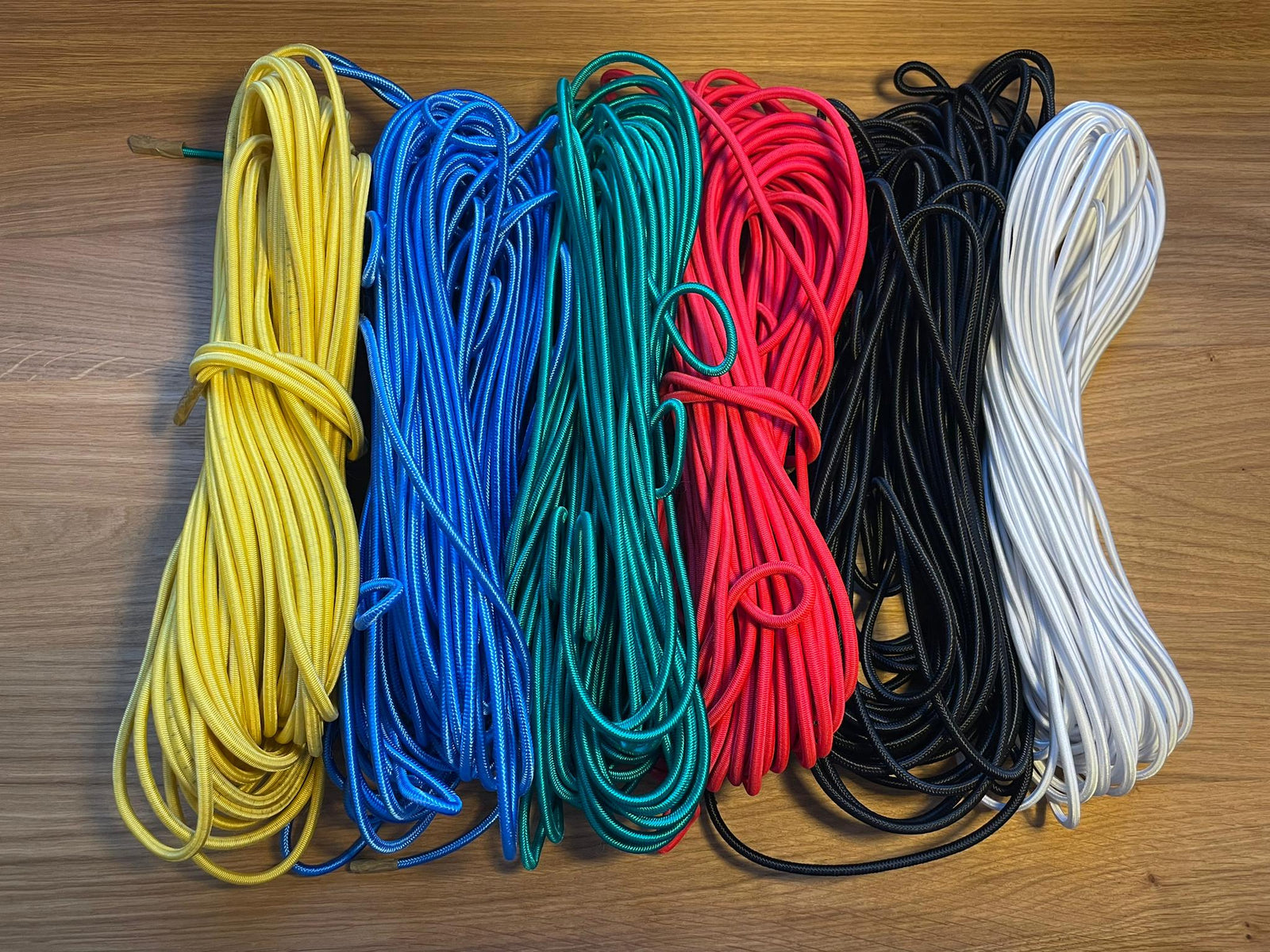 Sechs aufgerollte Leinen - gelb, blau, grün, rot, schwarz und weiß - von Rapid Surf Shop's Magnetleash mit Gummizug sind auf einer Holzfläche aufgereiht. Perfekt für Magnet-Leash- oder Quick-Release-Systeme; organisiert und verhedderungsfrei.