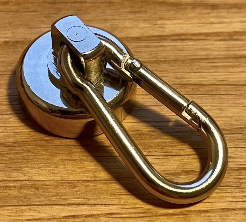 Ein metallisch glänzender, runder Neodym-Magnet (Ersatzteil) von Rapid Surf Shop mit silberner und goldener Oberfläche ist an einem goldenen Karabiner befestigt und ruht auf einer Holzoberfläche.