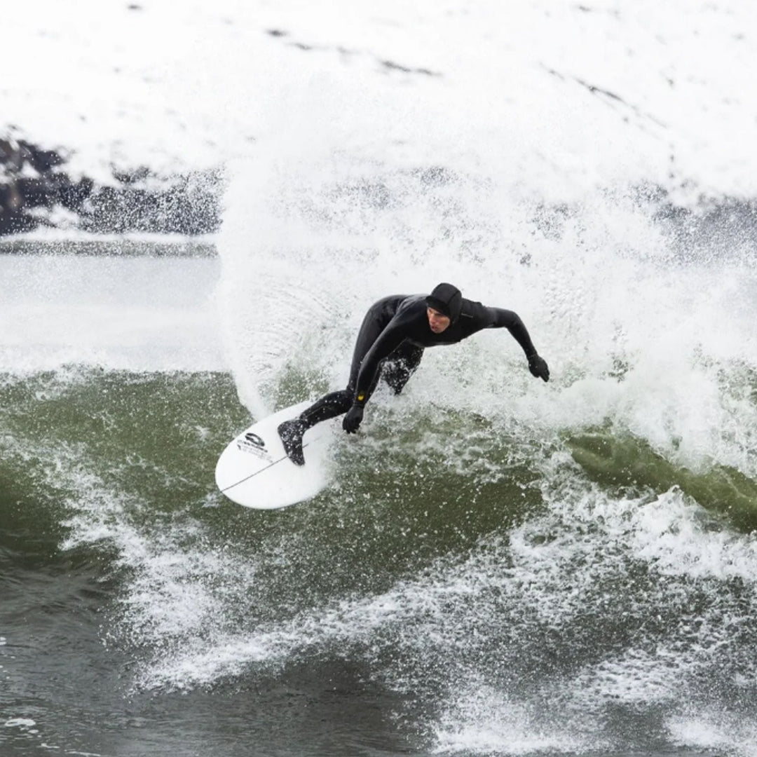 Ein Surfer in einem schwarzen Superstretch Neoprenanzug und einer MDNS Neopren Haube Prime 3mm Full Hood reitet eine Welle auf einem weißen Surfbrett, das Wasser spritzt umher, im Hintergrund sind weitere Wellen und ein bewölkter Himmel zu sehen.