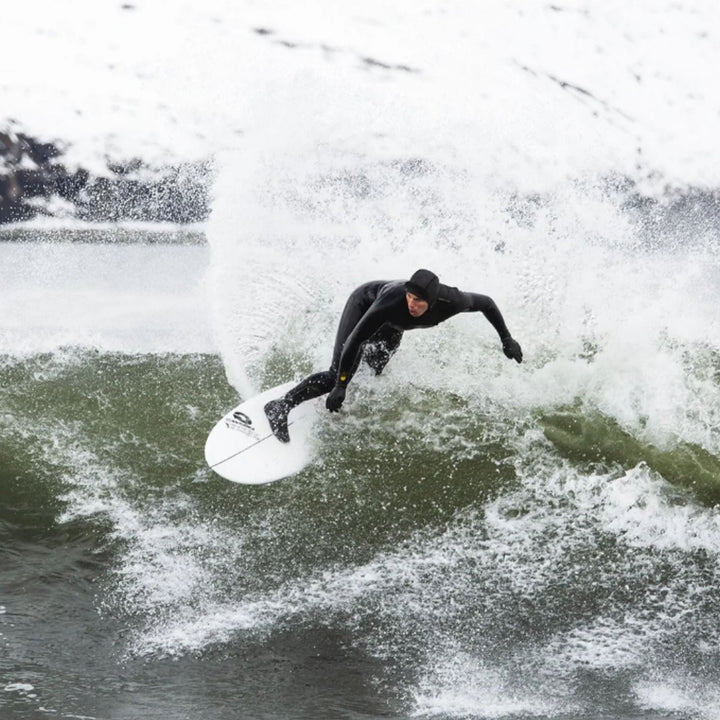 Ein Surfer in einem schwarzen Superstretch Neoprenanzug und einer MDNS Neopren Haube Prime 3mm Full Hood reitet eine Welle auf einem weißen Surfbrett, das Wasser spritzt umher, im Hintergrund sind weitere Wellen und ein bewölkter Himmel zu sehen.