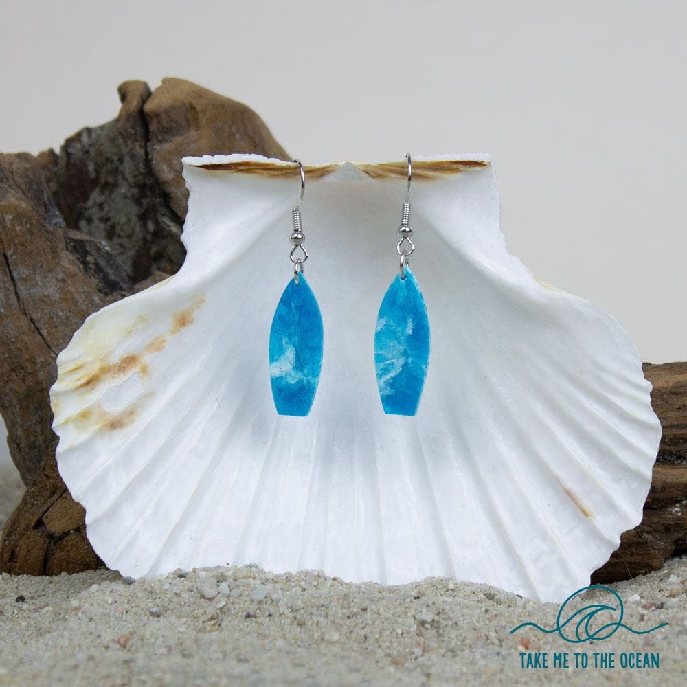 Ein Paar Surfboard Ohrringe handgefertigt von Takemetotheocean hängen an einer weißen Muschel, mit Treibholz und Sand dahinter. Das Takemetotheocean-Wellenlogo und "TAKE ME TO THE OCEAN" erscheinen in der unteren rechten Ecke.