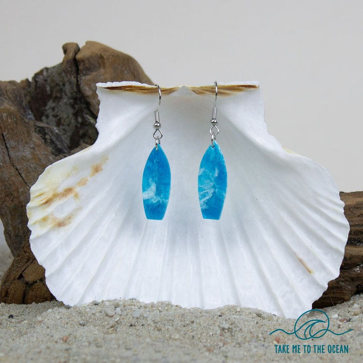 Ein Paar Surfboard Ohrringe handgefertigt von Takemetotheocean hängen an einer weißen Muschel, mit Treibholz und Sand dahinter. Das Takemetotheocean-Wellenlogo und "TAKE ME TO THE OCEAN" erscheinen in der unteren rechten Ecke.