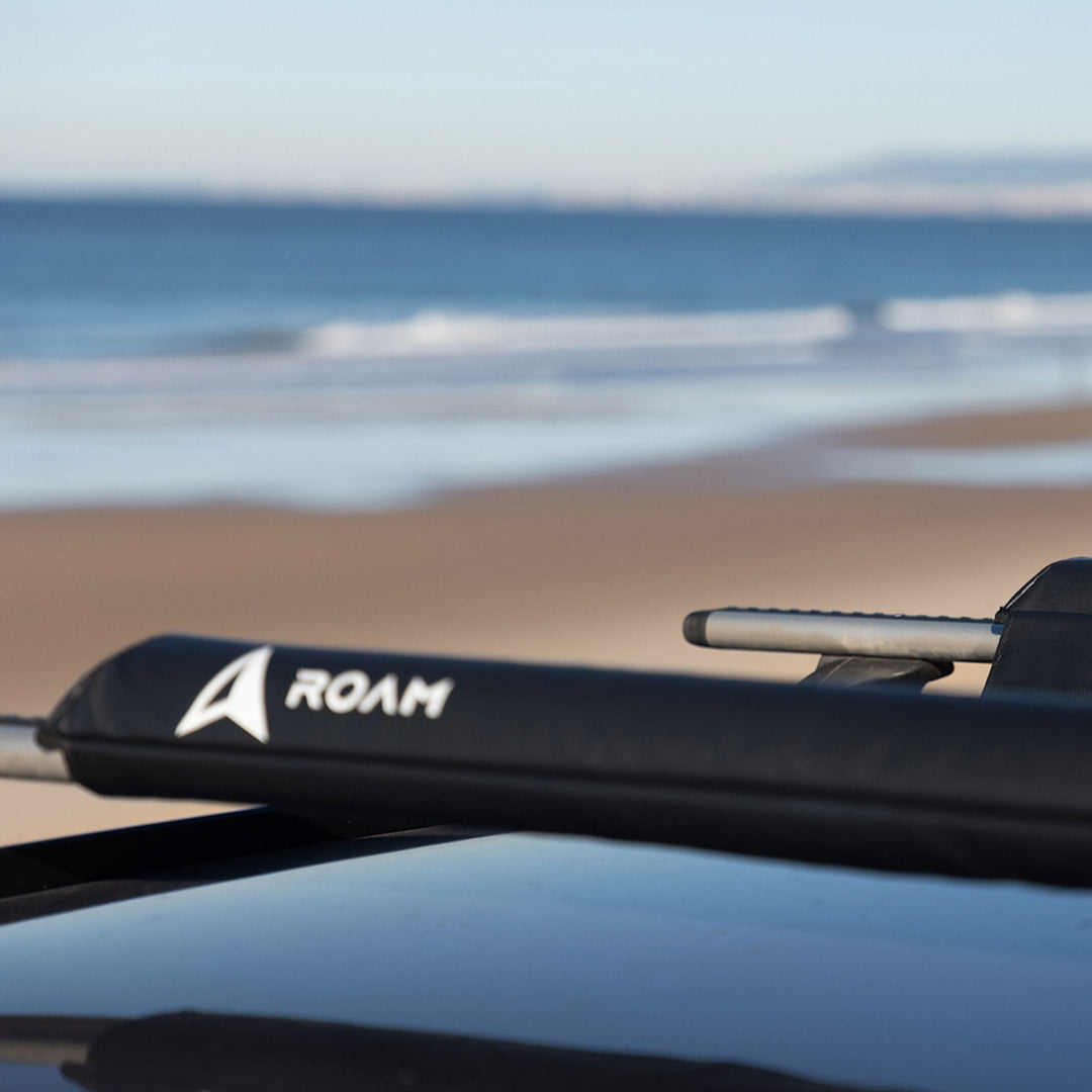 Eine Nahaufnahme eines Roam ROAM Dachauflage Surfboard Flach Polster Pad auf einem Fahrzeug, mit einem Sandstrand und Meereswellen im Hintergrund unter einem klaren Himmel.