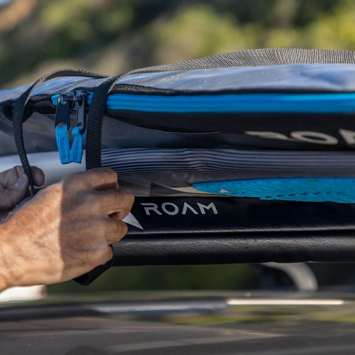 Eine Nahaufnahme von Händen, die ein Surfbrett auf einem Roam Dachauflage Surfboard Flach Polster Pad mit Riemen befestigen; im Hintergrund ist das unscharfe Grün im Freien zu sehen.