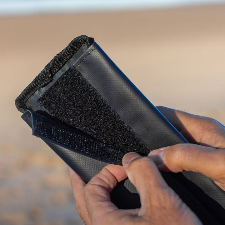 Eine Nahaufnahme von zwei Händen, die ein ROAM Dachauflage Surfboard Flach Polster Pad von Roam - mit einem sicheren Klettverschluss auf strapazierfähigem, strukturiertem Material - vor einem unscharfen Outdoor-Hintergrund befestigen.