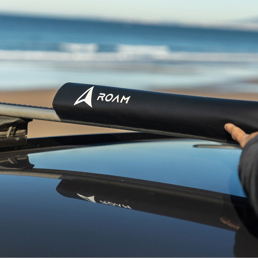 Eine Person justiert ein schwarzes ROAM Dachauflage Surfboard Rund Polster Pad auf einem Autodach, mit einem Sandstrand und Meereswellen im Hintergrund.