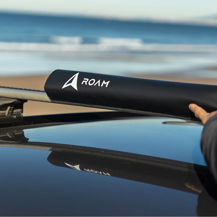 Eine Person justiert ein schwarzes ROAM Dachauflage Surfboard Rund Polster Pad auf einem Autodach, mit einem Sandstrand und Meereswellen im Hintergrund.