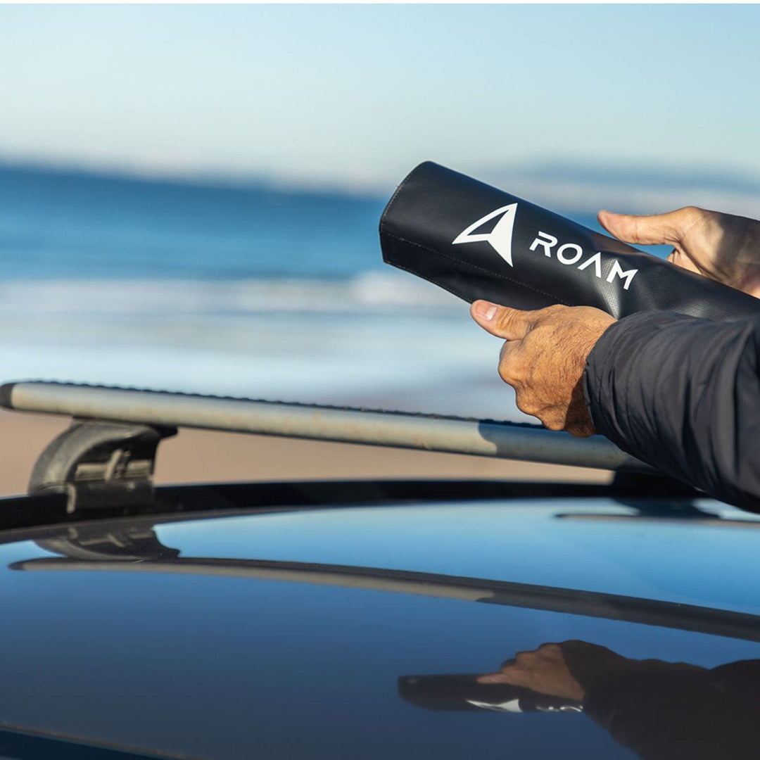 Eine Person steht an einem Auto mit Roam Dachauflage Surfboard Rund Polster Pads auf dem Dachgepäckträger und hält ein schwarzes zylindrisches Objekt mit der Aufschrift ROAM in der Hand, im Hintergrund das Meer.