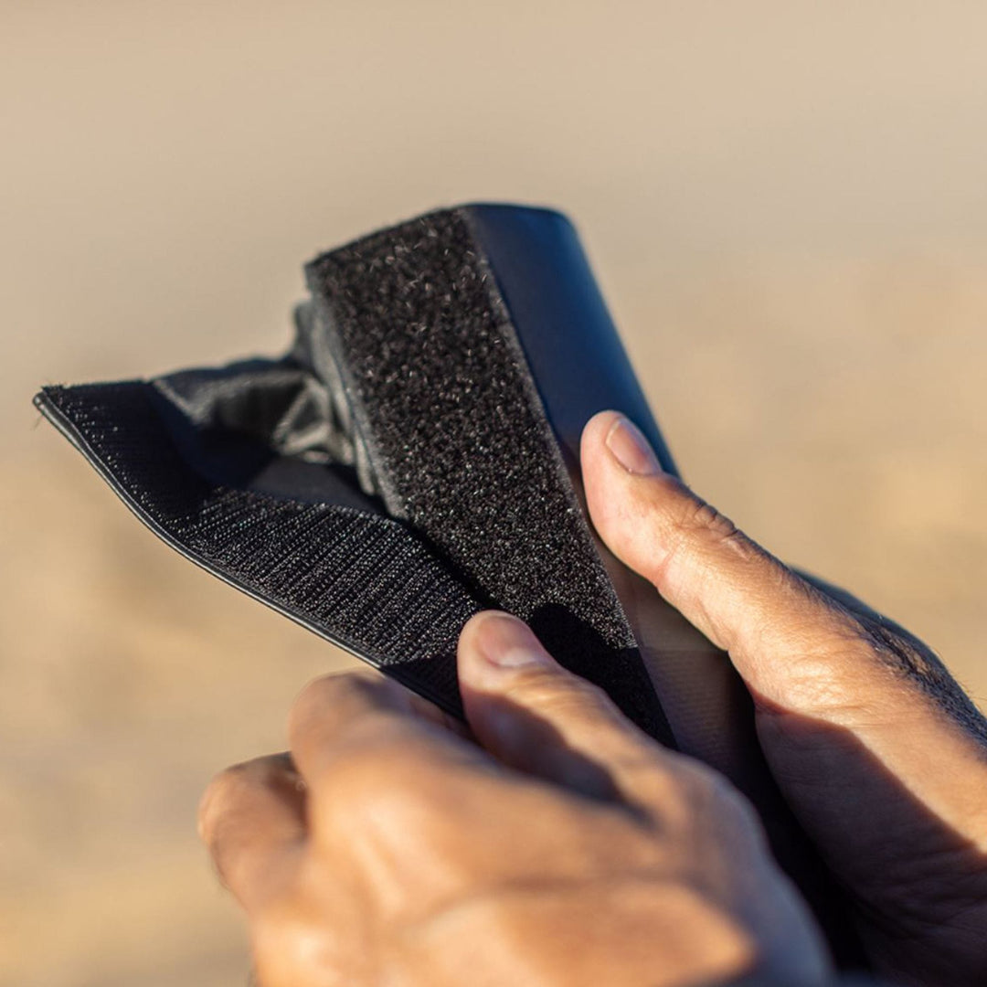 Die Hände einer Person befestigen einen schwarzen Klettverschlussriemen um ein ROAM Dachauflage Surfboard Rund Polster Pad von Roam, mit unscharfem Hintergrund.