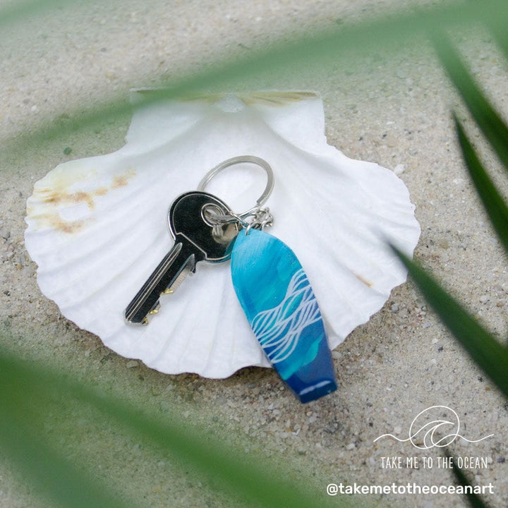 Ein Metallschlüssel und ein blauer Surfboard-Schlüsselanhänger handgefertigt von Takemetotheocean mit einem weißen Wellendesign ruhen auf einer weißen Muschel im Sand. Unscharfe grüne Blätter umrahmen die Szene, mit dem @takemetotheoceanart Logo in der Ecke.