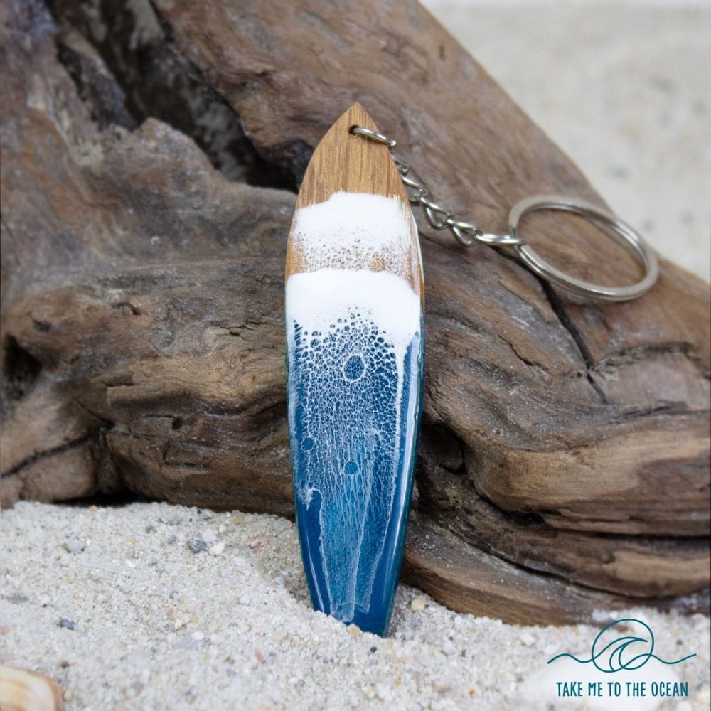 Der Surfboard Schlüsselanhänger handgefertigt von Takemetotheocean zeigt blaue und weiße Resin Ocean Art, die an Wellen erinnert. Er liegt auf Sand und Treibholz, das Markenlogo ist unten rechts sichtbar.