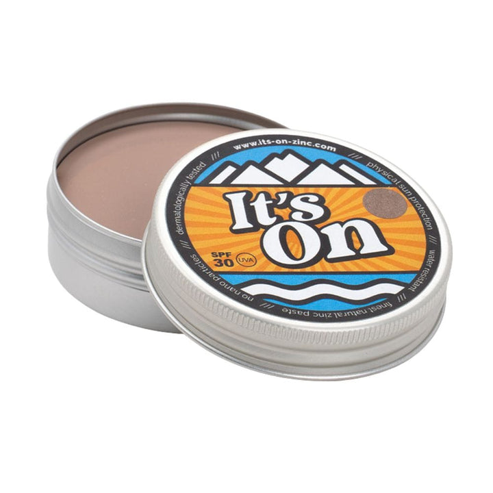 Eine runde Metalldose mit teilweise geöffnetem Deckel enthüllt eine bräunliche Creme im Inneren. Auf dem Etikett steht It&