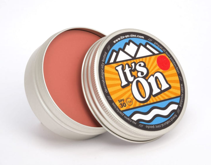 Eine runde Metalldose, teilweise geöffnet, enthüllt einen rötlich-braunen natürlichen Inhaltsstoffe Balm. Auf dem Deckel sind Berge, Wellen, eine rote Sonne und der Text "It&