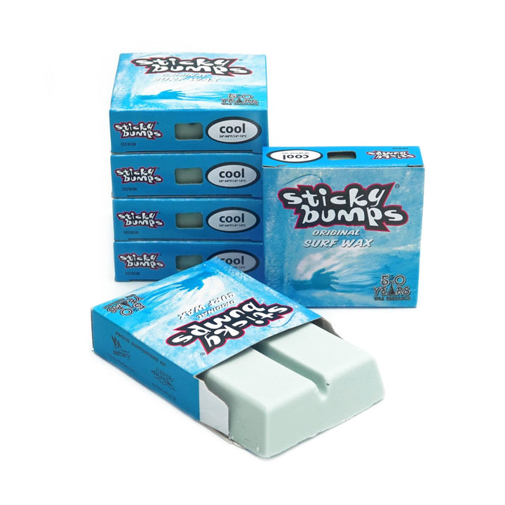 Fünf Schachteln Sticky Bumps - Surfwachs von Sticky Bumps sind ausgestellt, vier gestapelt und eine vorne offen mit zwei Stücken hellblauem, biologisch abbaubarem Wachs. Die Verpackung ist blau mit weißem und rotem Text.