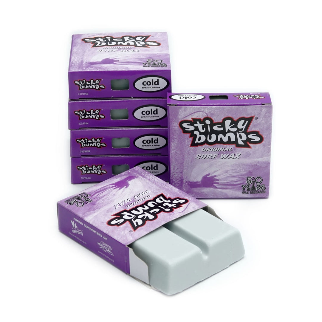 Fünf Schachteln Sticky Bumps - Surfwachs (kalt), lila-weiße Verpackung, links gestapelt; eine offene Schachtel vorne zeigt zwei hellgraue Balken. Biologisch abbaubar für optimalen Surf Grip.