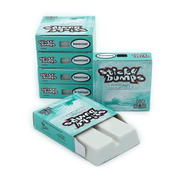 Mehrere Schachteln Sticky Bumps - Surfwachs von Sticky Bumps mit türkisfarbener Verpackung und der Aufschrift "Basecoat for ideal Surf Grip" sind übereinander gestapelt; in einer geöffneten Schachtel befindet sich ein weißer Riegel mit biologisch abbaubarem Wachs.