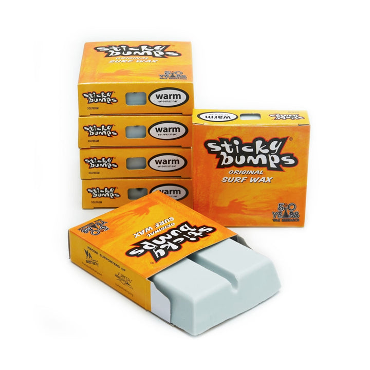 Fünf Schachteln Sticky Bumps - Surfwachs von Sticky Bumps mit der Aufschrift "warm" stehen neben einer offenen Schachtel mit zwei weißen Surfwachsriegeln. Die orangefarbene Verpackung hebt "Top Surf Grip" in fetter gelber und weißer Schrift hervor.
