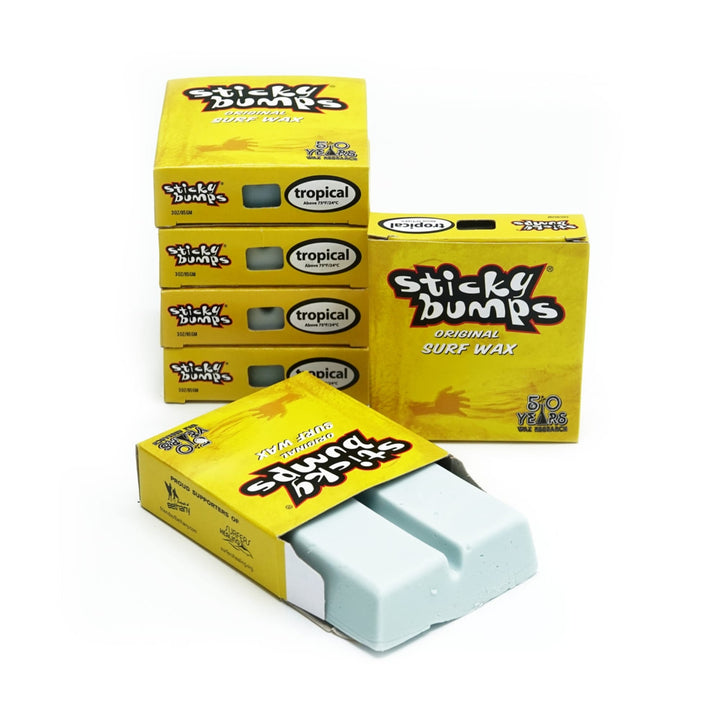 Fünf Schachteln Sticky Bumps - Surfwachs sind ausgestellt, eine davon geöffnet, um zwei hellblaue Stangen zum Vorschein zu bringen. Auf der gelben Verpackung steht "tropical" und "50 years" und verspricht Surf Grip für dein Board. Marke: Sticky Bumps.