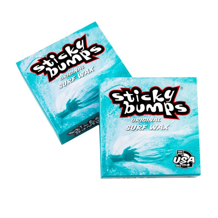 Abgebildet sind zwei blaue Schachteln Sticky Bumps - Surfwachs von Sticky Bumps, mit einer Hand unter Wasser und einem auffälligen Branding. Die Verpackung hebt seine USA Herkunft und dass es biologisch abbaubar-perfekt für Surf Grip ist.