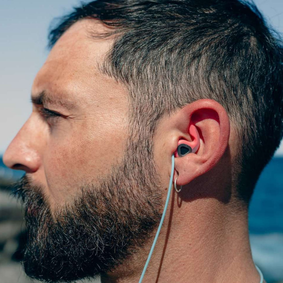 Nahaufnahme einer bärtigen Person, die Surf Ears 4.0 von Surf Ears und einen kleinen Ohrring trägt und im Freien am Wasser steht.