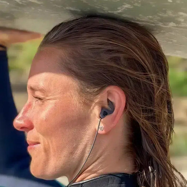 Eine Person mit nassen Haaren, die Surf Ears 4.0 von Surf Ears trägt, hält ein Surfbrett über Kopf im Freien im Sonnenlicht, wobei das Grün im Hintergrund verschwimmt.