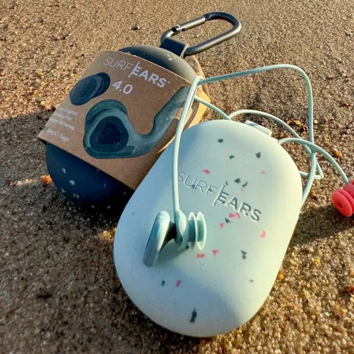 Zwei Surf Ears 4.0 Etuis von Surf Ears liegen im Sand: das eine ist dunkel mit Pappetikett und Karabiner, das andere hell mit gesprenkeltem Design und angebrachten blauen Gehörschutz-Surfen Ohrstöpseln.